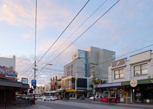 Urban streetscape showing a commercial main street with low-rise shopfronts, restaurants and cafés, overhead utility and tram wires, street signage, and a taller contemporary concrete-and-glass building in the background. The scene is photographed from the street at dusk with soft pastel light.