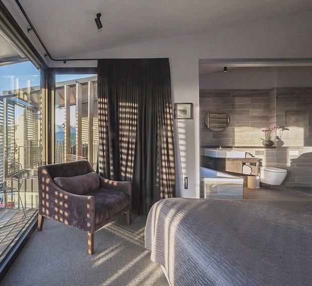 A modern bedroom with floor-to-ceiling glazing opening onto a balcony, a dark upholstered armchair, textured grey bedspread, and dark curtains. An open ensuite bathroom is visible directly beside the bed, with grey horizontal wall tiles, a floating timber vanity, white basin, and wall-hung toilet. Strong afternoon sunlight creates patterned shadows across the room.