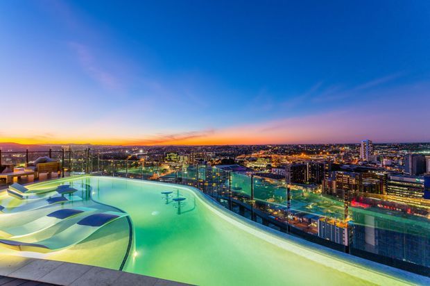 A rooftop infinity pool at dusk, with sculptural curved edges and shallow lounging areas in the foreground. Glass balustrades frame expansive views over a brightly lit urban cityscape beneath a blue-to-orange sunset sky.