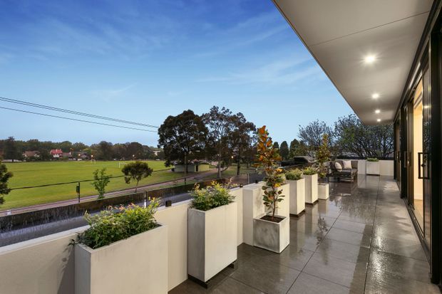 A spacious covered outdoor terrace or balcony runs along the side of a contemporary building, furnished with outdoor seating and lined with large rectangular planter boxes. Grey tiled paving, a white rendered balustrade, recessed ceiling lights, and dark-framed glazing create a clean modern setting. Beyond the terrace is an open green suburban landscape with trees and low-rise homes.