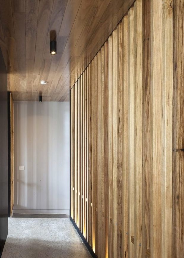 A narrow interior hallway with a warm timber-lined ceiling and a prominent vertical timber slat feature wall or screen along the right side. The corridor leads to a plain light-coloured door, with a polished concrete or terrazzo-like floor and discreet cylindrical recessed and surface-mounted lighting. The design is pared-back, natural and contemporary, with strong linear detailing and warm wood tones.