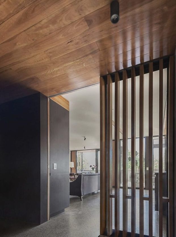 Interior view looking through a partially open doorway into a refined living area. The foreground features a warm timber-lined ceiling and a dark timber vertical slatted screen, with dark painted wall and door surfaces. Beyond is a spacious living room with grey seating, polished concrete flooring, large windows, and restrained contemporary furnishings. A black cylindrical ceiling light is visible overhead.