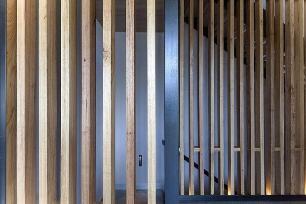Close-up interior view of a vertical timber-slat screen or balustrade, with varied natural wood tones and narrow openings revealing a dark blue-grey wall, doorway, and stair structure behind. The repeating slats create a strong linear architectural feature and partially screen the circulation area.