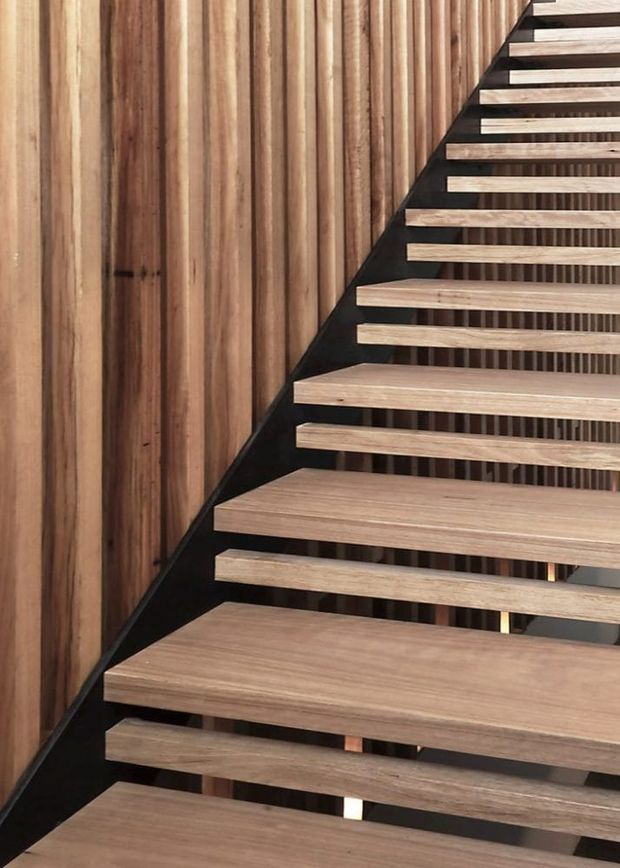 Close-up view of a contemporary open-riser staircase with light timber treads supported by a dark metal structure. The staircase rises alongside a vertical timber slat feature wall, creating a strong linear rhythm and warm natural-material palette.