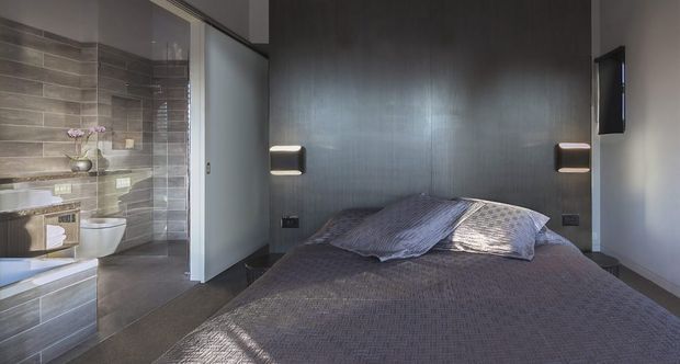 Compact contemporary bedroom with a low-profile bed facing a dark vertically textured feature wall, integrated bedside wall lights, and a partially open ensuite bathroom visible at left. The ensuite includes grey-brown wall tiles, a wall-mounted toilet, floating vanity cabinetry, and a glass shower enclosure.