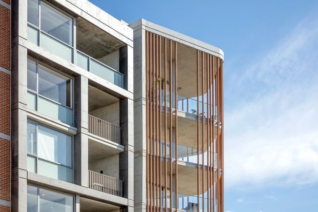 Exterior architectural photograph of a contemporary multi-storey apartment building. The facade combines exposed concrete balcony slabs and columns, extensive glazing, brickwork at the edge, and a prominent rounded corner screened by slender vertical timber-toned fins. Multiple balconies and recessed outdoor spaces are visible against a clear sky.