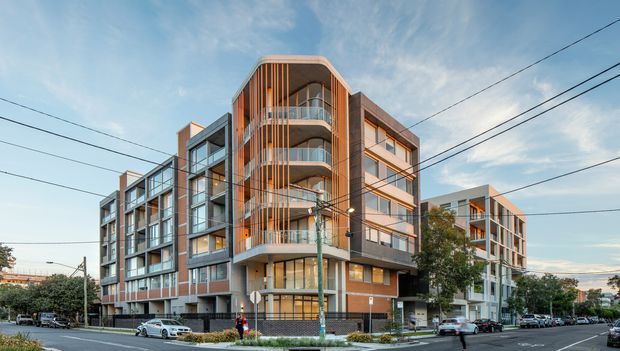 Street-level exterior view of a contemporary multi-storey apartment building on an urban corner site. The building has articulated wings, extensive glazing, rounded projecting balconies, vertical timber-toned screening, exposed balcony edges, and a mix of dark and light facade panels. Streets, parked vehicles, utility wires, and mature street trees surround the development.