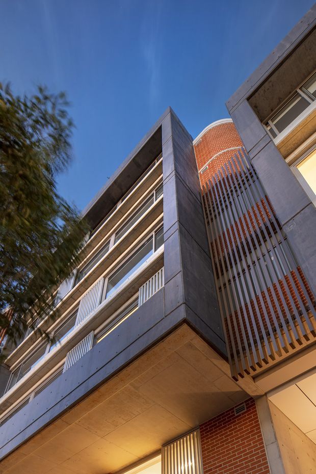 Upward-looking architectural photograph of a multi-storey contemporary building. The facade combines dark grey concrete or stone-like panels, horizontal balconies with light railings, exposed warm red brick, and a distinctive curved vertical slatted screen around an upper-level balcony. Warm lighting is visible beneath the projecting structure, with blue sky and a blurred tree in the background.