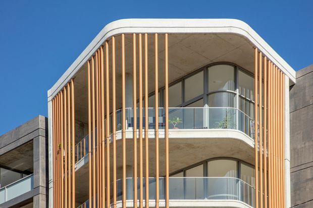 Exterior architectural photograph of a contemporary multi-storey building with a rounded, projecting facade. Curved balconies with clear glass balustrades are screened by closely spaced vertical timber-toned fins, framed by exposed pale concrete soffits and side walls. Arched and curved glazing reinforces the soft geometric form against a clear blue sky.