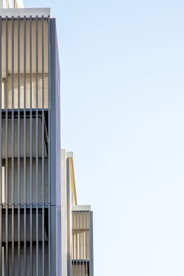 Close-up exterior view of a contemporary multi-storey building facade, featuring repetitive vertical dark metal fins screening recessed balconies or windows. Pale rendered or concrete wall surfaces contrast with the dark vertical elements, while a large expanse of clear blue sky occupies the right side of the composition.