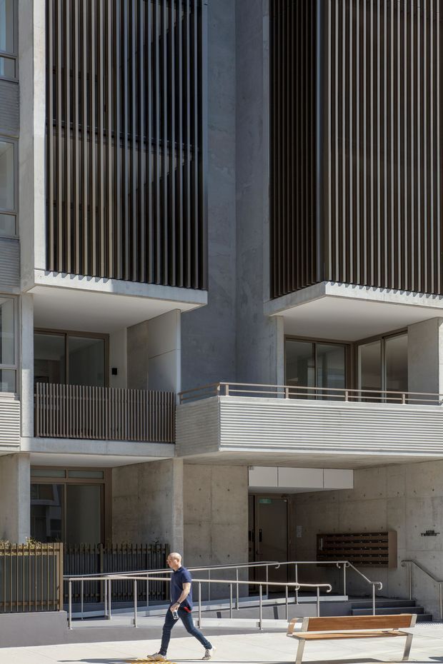 Street-level view of a contemporary multi-storey apartment building facade. The building features exposed concrete columns and soffits, large glazed openings, dark vertical timber or metal privacy screens, projecting balconies with horizontal metal balustrades, and an accessible ramp and entry stair. A pedestrian and bench are visible in the foreground.
