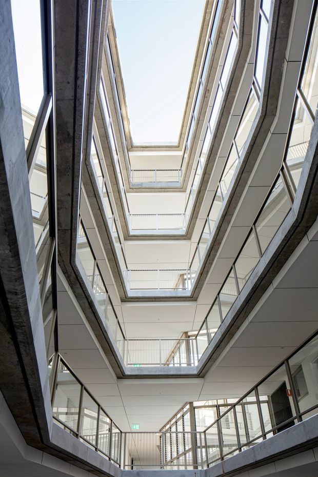 Vertical interior view looking upward through a multi-storey atrium or circulation void. Repeated gallery levels with exposed concrete slab edges, pale panelled ceilings, glass and metal-framed balustrades, and extensive glazing surround a central open shaft topped by a skylight. The image has a clean, contemporary institutional or commercial character and strong vertical perspective.