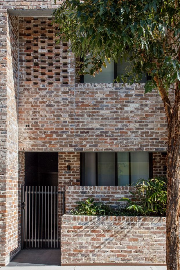 A close exterior view of a multi-level brick residential facade. The building features exposed recycled-style brickwork, a perforated brick screen, dark-framed windows, a black metal pedestrian gate, and a raised brick planter with greenery. A mature tree partially screens the facade. The composition highlights textured masonry and layered courtyard-like frontage.