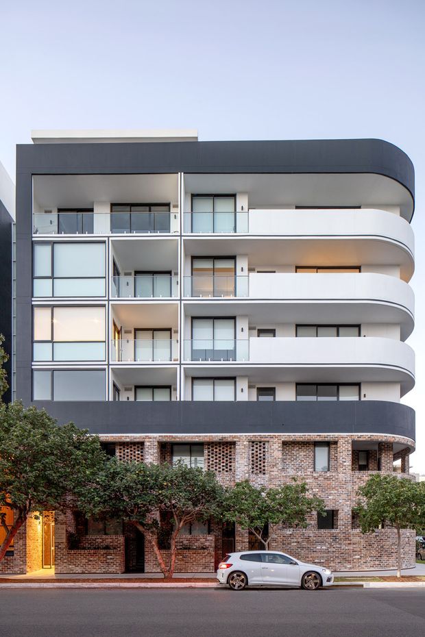 Street-facing photograph of a contemporary multi-storey apartment building. The facade combines dark charcoal cladding with smooth white curved balcony bands, extensive glazing, and a lower level clad in patterned exposed brick. Mature street trees partially screen the ground floor, and a parked car is visible on the road in front.