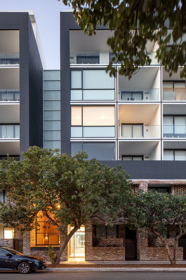Street-facing view of a contemporary multi-storey apartment building, with a dark charcoal facade, exposed brick lower level, balconies, frosted and clear glazed panels, and regular box-like window bays. Mature street trees partially screen the frontage, with a warmly illuminated entry at ground level and a parked car along the curb.