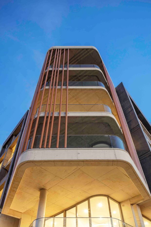 Low-angle exterior photograph of a multi-storey contemporary building with stacked rounded balconies, exposed concrete soffits and columns, curved glass balustrades, and vertical terracotta-coloured metal fins. The warm-toned facade contrasts strongly with the clear blue sky.