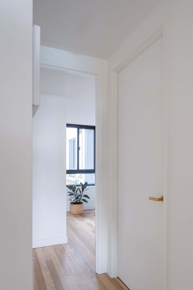 A bright, narrow residential hallway viewed toward an adjoining room. The space has white painted walls and ceiling, decorative white door and architrave mouldings, a closed white interior door with a small brass-gold lever handle, and light timber flooring. Through the open doorway, a black-framed window, pale timber floor, and a potted plant are visible.