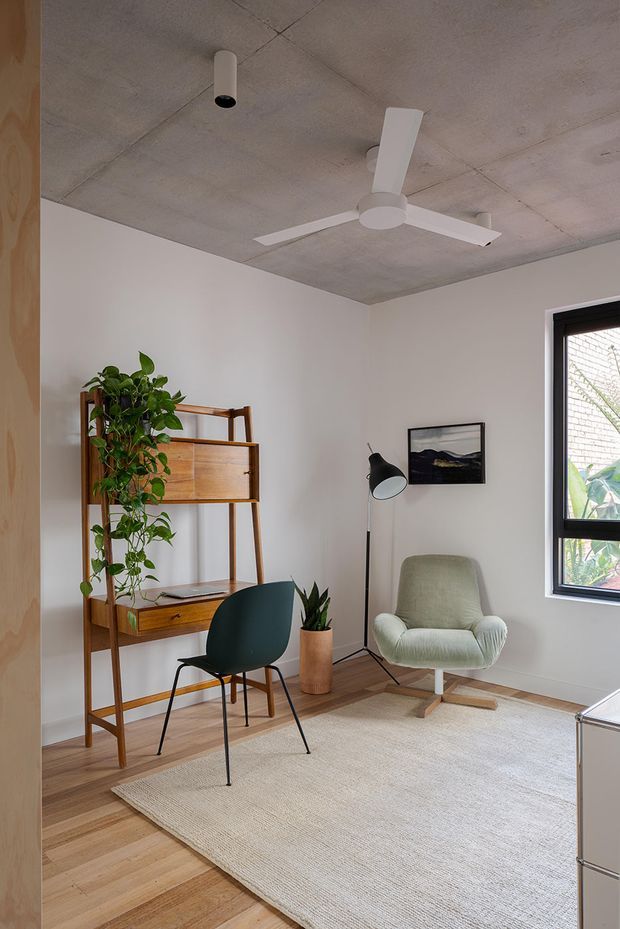 A bright, compact home office or study with white walls, a raw concrete ceiling, timber flooring, and a large neutral woven rug. The room contains a mid-century-inspired timber shelving and desk unit, dark green desk chair, pale upholstered lounge chair, black floor lamp, indoor plants, framed artwork, and a black-framed window. A ceiling fan and minimalist cylindrical light add contemporary functional details.