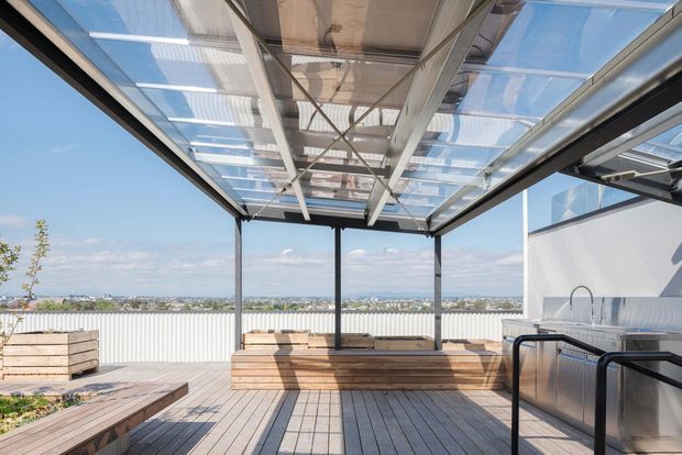 A bright rooftop outdoor entertaining area with a timber deck, built-in timber bench seating, raised planter boxes, and a compact outdoor kitchen or bar at the right. The space is sheltered by a transparent pitched canopy supported by dark metal framing, with expansive views across the surrounding suburb. The design is contemporary and open-air, with strong indoor-outdoor and rooftop terrace characteristics.