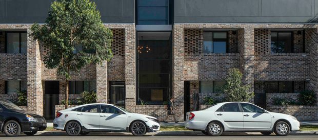 Street-level exterior view of a contemporary multi-storey brick apartment or townhouse building. The facade combines dark brickwork, deep rectangular window openings, recessed balconies or openings, and a central glazed entrance with a tall vertical window section. Mature trees and parked cars line the street in front.