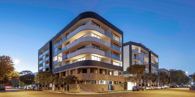 Twilight exterior view of a contemporary multi-storey apartment building on a street corner. The building features curved projecting balconies, dark framed glazing, warm interior lighting, a rounded dark roof element, and a textured brick-clad podium. Trees, parked cars, and surrounding streets are visible in the foreground and background.
