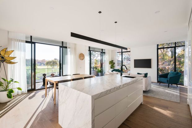 Bright open-plan contemporary kitchen and living area with a large white marble-look island, integrated white cabinetry, timber flooring, floor-to-ceiling black-framed glazing, and a minimalist linear pendant light. A dining table and living seating area are visible beyond the island, with abundant natural light and leafy views outside.