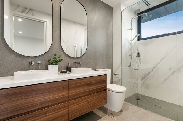 Contemporary bathroom featuring a double floating timber vanity with white vessel basins, rounded black-framed mirrors, a wall-mounted tap set, toilet, and frameless glass walk-in shower. Large marble-look wall tiles and a textured grey feature wall create a refined, neutral finish, with a high horizontal window providing natural light.