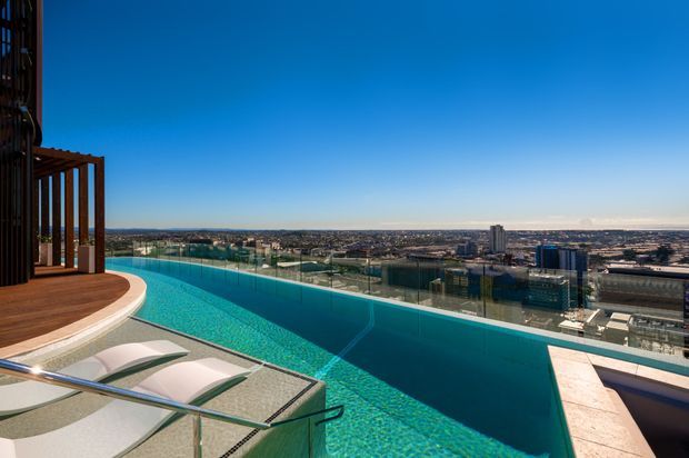 Elevated rooftop or high-level infinity pool terrace with expansive city and waterfront views. The pool has turquoise mosaic tiling, a glass balustrade, timber decking, curved built-in steps or loungers, and a dark timber pergola structure at the left. Bright clear conditions create a contemporary, luxurious outdoor setting.