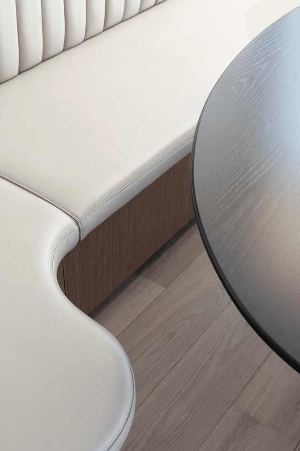 Close-up interior detail showing a curved cream upholstered banquette with vertical channel tufting, a dark wood-toned base, a round dark tabletop, and light timber flooring. The composition highlights refined built-in seating and material contrasts.