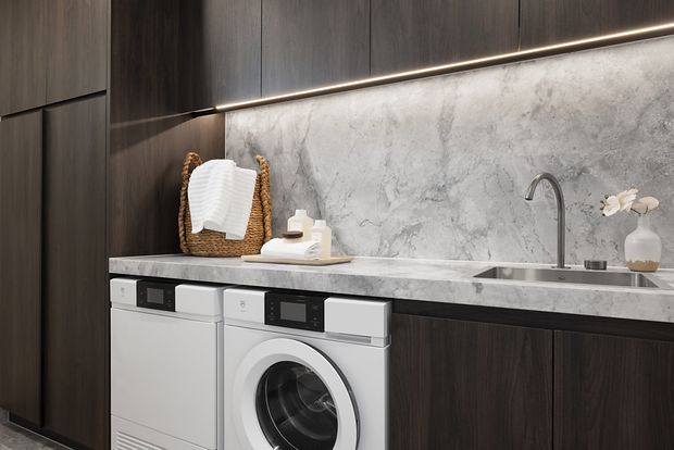Contemporary laundry room with dark wood cabinetry, a grey marble-look benchtop and splashback, integrated linear lighting, front-loading white washing machine and dryer, and a stainless steel gooseneck sink tap. A woven basket, folded towels, and small decorative accessories sit on the counter.