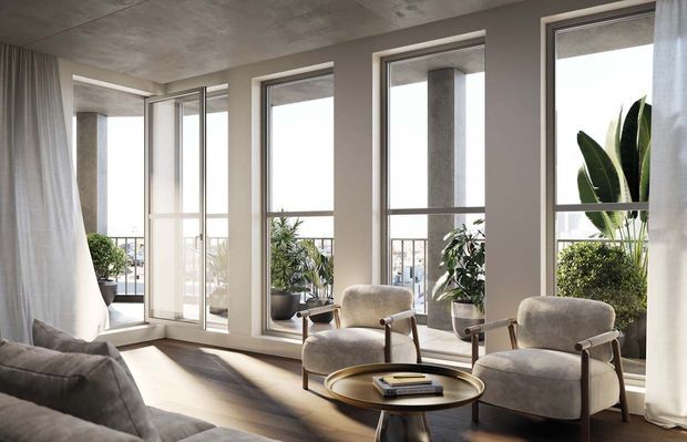 Bright contemporary living room with floor-to-ceiling windows and a glazed balcony or terrace beyond. The room features a concrete ceiling, pale walls, light timber flooring, sheer curtains, a grey sofa, two upholstered lounge chairs, and a round coffee table. Potted plants and concrete structural columns are visible outside the glazing, with abundant natural light creating an airy, refined atmosphere.