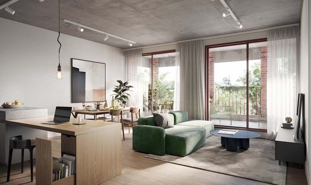 A contemporary open-plan apartment interior combining a kitchen, dining area, living room and compact workspace. The room features a pale timber kitchen island and cabinetry, concrete ceiling, light flooring, large glazed balcony doors with sheer curtains, a green upholstered sofa, blue coffee table and track lighting. The space is bright, refined and minimally styled, with exposed brick visible around the exterior glazing.