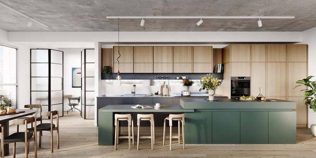 A bright open-plan kitchen and dining area with a large dark green island, light timber cabinetry, white tiled splashback, integrated appliances, timber flooring, and a raw concrete ceiling with track lighting. A black-framed glazed partition separates the dining area from an adjacent space.
