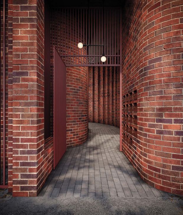 A narrow architectural passageway framed by curved and straight red brick walls, with a dark red metal security gate and vertical bars. Grey rectangular pavers lead through the entry, while spherical wall lights and brick detailing add visual interest. The image emphasizes the textured masonry and enclosed urban entrance.