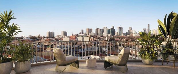 A rooftop or high-level outdoor terrace overlooking a dense urban skyline. Two woven lounge chairs and a small round table sit on a paved terrace, framed by large potted tropical plants and a pale metal balustrade. The scene is bright, open, and contemporary with expansive city views.