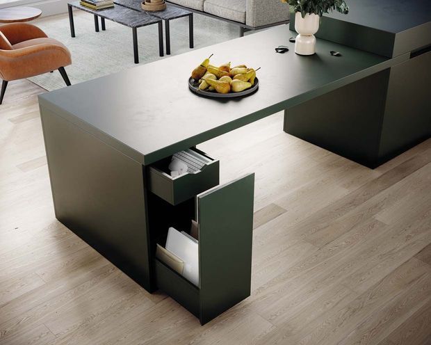 A contemporary open-plan kitchen and living area featuring a dark green island with a light stone benchtop, integrated drawers and storage, and pale timber flooring. A lounge setting with a sofa, coffee table and curved armchairs is visible in the background.