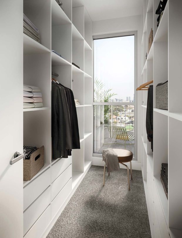 A narrow, bright walk-in wardrobe with full-height white built-in shelving, hanging rails, drawers, folded clothing and woven storage baskets. A large window at the end overlooks greenery and nearby buildings, while a small upholstered stool sits on a textured grey carpet.