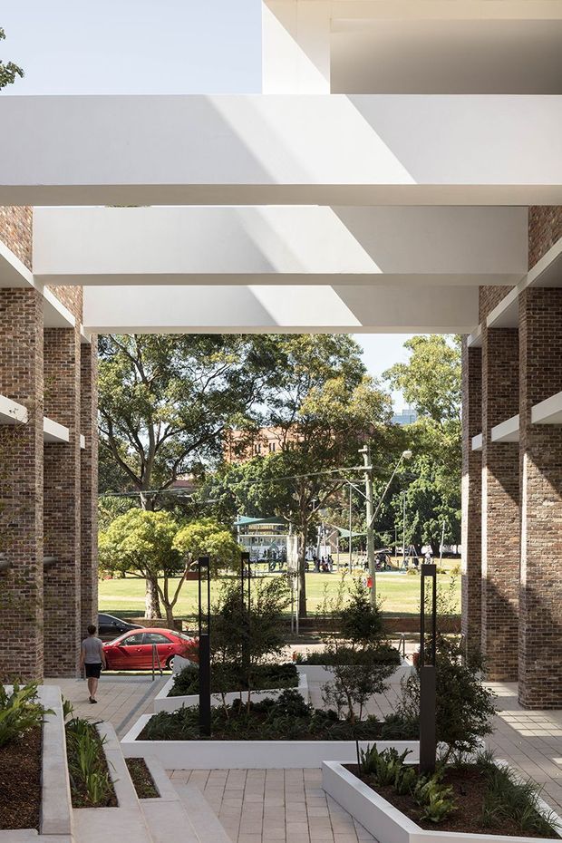 A covered, open-air architectural passage or courtyard framed by exposed red-brick walls and broad white concrete beams. Rectangular skylight openings cast strong geometric sunlight and shadow across the overhead structure. Integrated white raised planters with low landscaping line a paved walkway, opening toward a leafy urban park and street beyond.