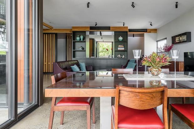 A refined open-plan living and dining area viewed from beside full-height glazing. The room features dark built-in shelving and cabinetry, warm timber detailing, leather lounge seating, a polished dark dining table, and mid-century-inspired timber dining chairs with red upholstered seats. Track lighting and large windows create a bright contemporary interior with views to greenery outside.
