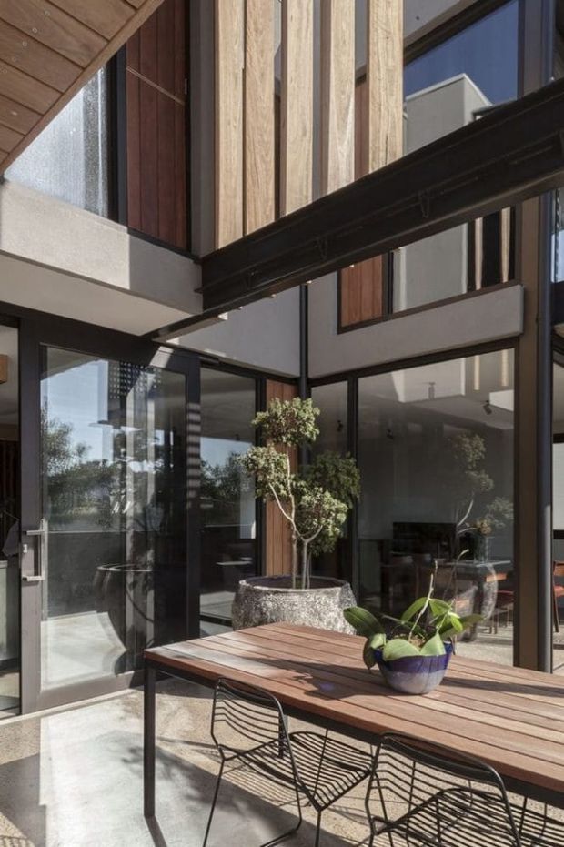 A sunlit modern courtyard or covered outdoor dining area within a multi-level home. The space features a timber dining table with black wire chairs, large black-framed glazed doors and windows, vertical timber cladding, exposed dark steel beams, concrete paving, and a raised planter with a small tree. Interior spaces are visible through the glazing.