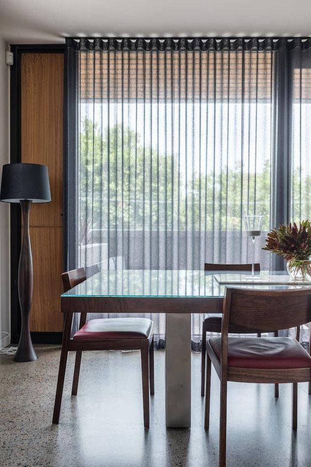 A refined dining area with a dark timber dining table and matching chairs, set in front of full-height glazing dressed with sheer charcoal curtains. A tall sculptural black floor lamp stands beside a built-in timber panel, while a floral arrangement adds a natural accent. The room has a polished, contemporary feel with strong daylight and views to greenery beyond the glass.