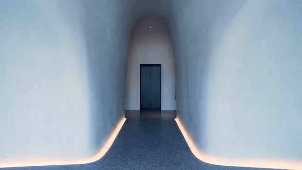 A minimalist, dimly lit architectural corridor with smooth pale walls curving seamlessly into the dark floor. Warm recessed floor-level lighting traces both sides toward a dark doorway at the end, creating a dramatic tunnel-like perspective.
