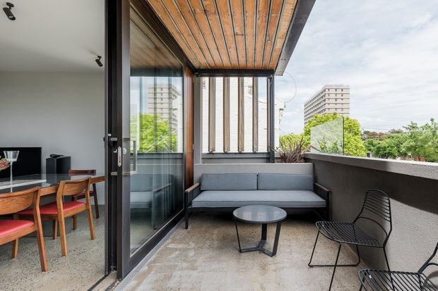 A contemporary apartment balcony terrace viewed from an open-plan interior, with a grey outdoor sofa, round coffee table and wire-frame chairs on a tiled floor. Timber-lined ceiling, dark-framed full-height glazing and a solid rendered balustrade define the compact outdoor space, while a dining area is visible inside and urban buildings appear beyond.