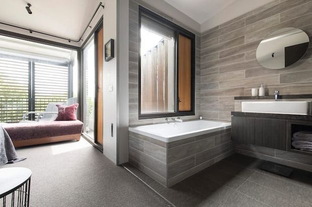 Contemporary bathroom with a built-in tiled bathtub, floating dark timber vanity, white rectangular basin, frameless oval mirror, and black-framed window. The bathroom opens directly into a carpeted bedroom or sitting area with a chaise lounge and louvred glazing.
