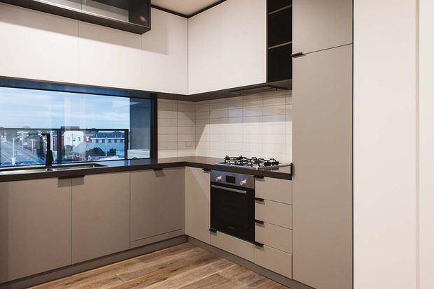 Compact contemporary L-shaped kitchen with white and taupe flat-panel cabinetry, dark benchtops, integrated black oven and gas cooktop, tiled splashback, timber-look flooring, and a large black-framed window overlooking an urban setting.