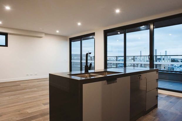 A bright, contemporary open-plan kitchen viewed from the living area, centred on a large dark kitchen island with an integrated sink and dishwasher. Light timber flooring, white walls, recessed ceiling lights, and extensive black-framed floor-to-ceiling glazing create an airy urban interior with city views beyond.