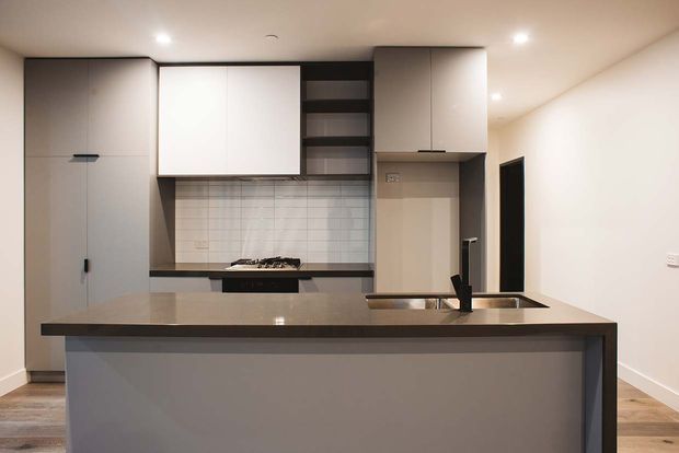 A contemporary compact kitchen viewed from the living area, featuring a large island with an integrated sink and dark tap, taupe-grey benchtop, matte grey cabinetry, white overhead cupboards, open shelving, and a light tiled splashback. Recessed ceiling lights and timber-look flooring complete the neutral, streamlined fit-out.