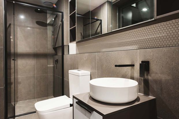 Compact contemporary bathroom featuring a frameless glass shower, wall-mounted black tapware, white vessel basin, toilet, large grey stone-look wall tiles, and a small hexagonal mosaic splashback beneath mirrored cabinetry.