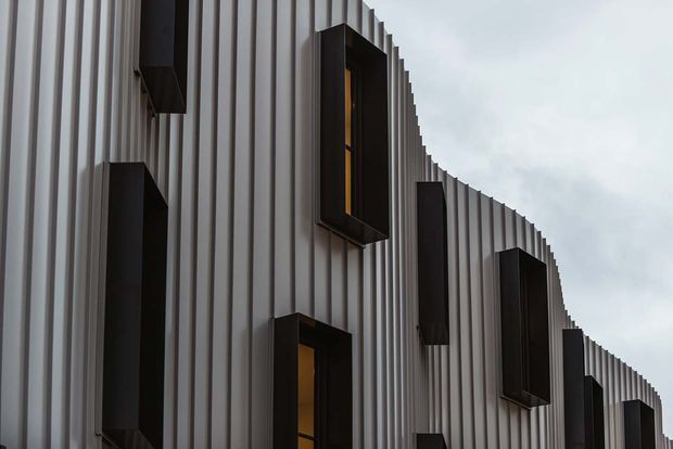 Close-up exterior view of a contemporary building facade with vertically ribbed light-grey cladding and a sweeping curved profile. Narrow black-framed window projections punctuate the facade, with warm interior light visible in some openings beneath an overcast sky.