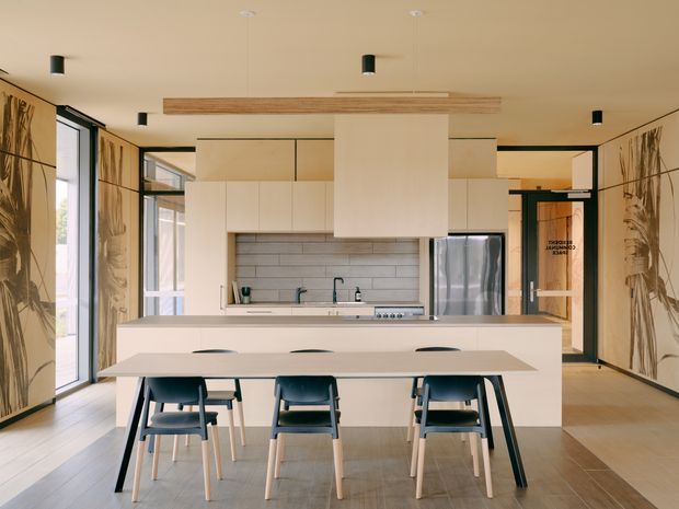 Bright contemporary open-plan kitchen and dining area with a large white island, integrated cabinetry, stainless-steel appliances, pale tiled splashback, and a long dining table with dark chairs. Timber-toned flooring, warm neutral surfaces, black-framed glazing, and decorative full-height wall panels create a refined, minimalist interior.
